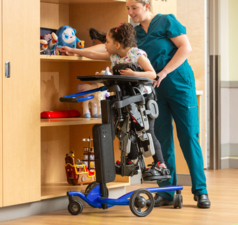 A girl in a blue Size 2 Rifton Stander reaches for a toy on a shelf with her caregiver helping