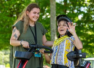 A girl riding on a Rifton Adaptive Tricycle waves and smiles with her caregiver.