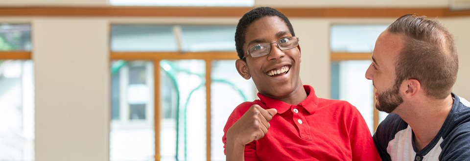 A young teenager in a red shirt grins at the camera as he poses with his caregiver.