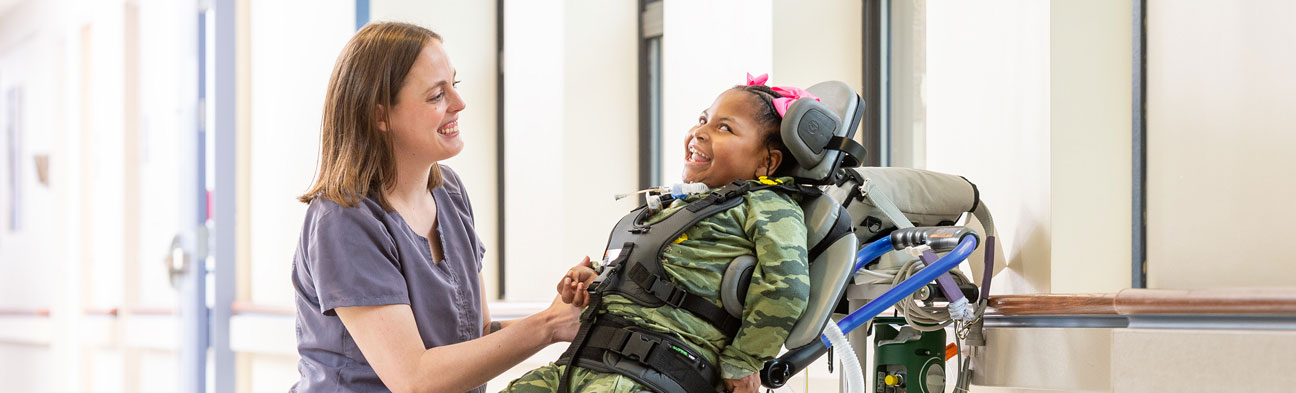 A girl standing in a Rifton Stander and smiling at her caretaker.