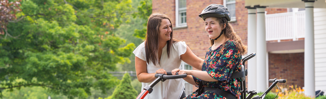 A young woman rides on a Rifton Adaptive Tricycle and smiles at her therapist.