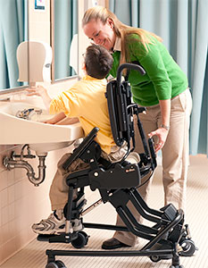 Adaptive seating equipment positions a boy for handwashing at a bathroom sink with assistance from a teacher's aide