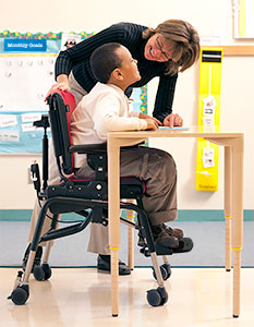 Adaptive seating positions this young boy at a school desk and recieve help from the teacher who is bending over him