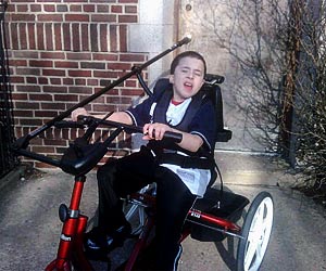 A young Carlos rides his red Rifton adaptive tricycle in front of a brick building