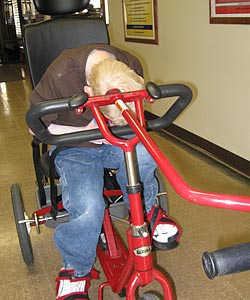 A young boy with muscle control issues hunched over on his red Rifton adaptive tricycle in the hallway of his elementary school because he lacks Rifton's chest prompt support system