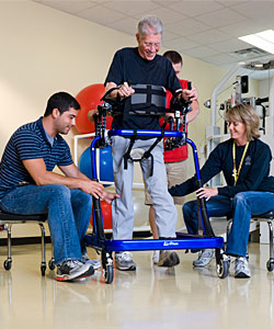 A man in a blue gait trainer practices ambulation aided by two PTs at a facility