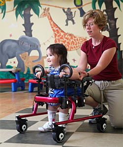 A therapist guides a special needs toddler in a red gait trainer in a playroom helping her perform early childhood intervention activities