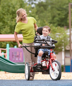 An early intervention therapist guides a young boy on a red Rifton tricyle through a park on a sunny day