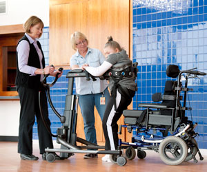 Two therapists assist a woman with disabilities to transfer from sitting to standing using the Rifton Transfer and Mobility Device