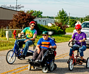 A young man and women with Extra Special People ride tricycles next to a boy in a wheelchair leading the Tour D’Oconee race while being tailgated by a police vehicle.