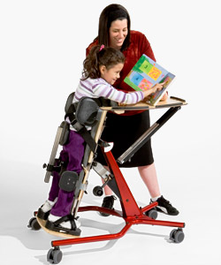 A child with special needs properly positioned in a Prone Stander points to a picture in a book as her caregiver stands smiling over her
