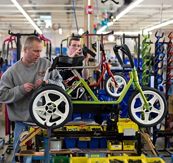 Colorful green and red tricycles on mounts are being adjusted by Rifton workers at the manufacturing facility
