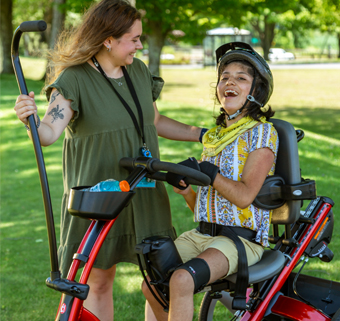 A girl rides on a Rifton Adaptive Tricycle and laughs happily with her caregiver.