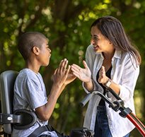 A boy sits on a trike while his caregiver smiles and claps with him.