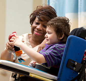 A young boy sits in his wheelchair as he practices reaching motor learning skills using a puppet that his therapist is giving to him.