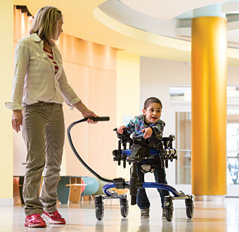 A therapist helps guide a boy with special needs in a gait trainer as he eagerly walks towards the music playing during his session