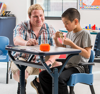 A boy sitting on a Rifton Compass Chair playing, with his caretaker sitting next to him