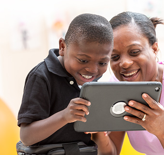 A boy in a Rifton Pacer looks at a communication device with his therapist.