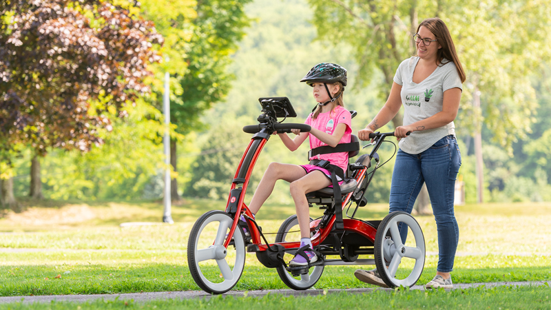 A girl pedals a red Rifton Adaptive Tricycle, while her caregiver steers from behind.