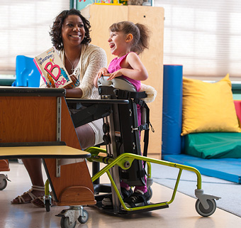 A girl stands in a Rifton Mobile Stander and reads a book with her caretaker.
