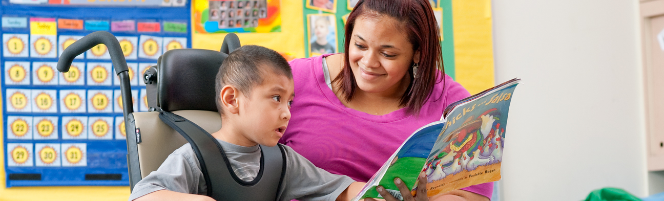 A boy sits in a Rifton Activity Chair while his caretaker reads a book to him.