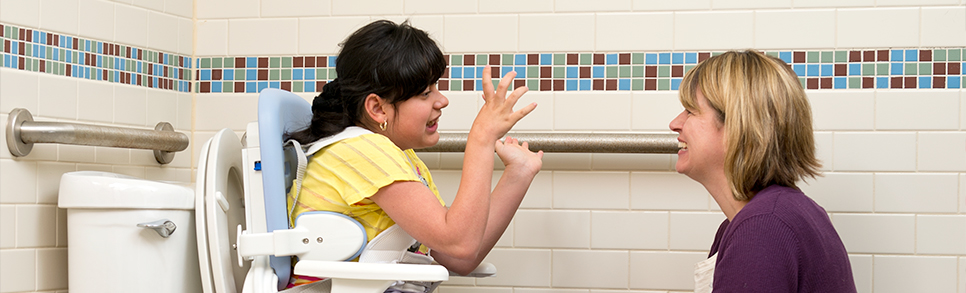 A girl sits on a Rifton HTS and talks with her caretaker.