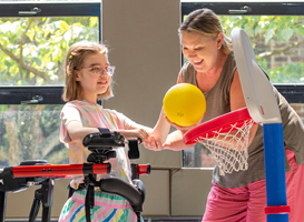 A girl stands in a Rifton Pacer in posterior position and plays basketball with her caretaker.