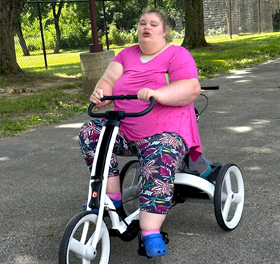 A young woman with disabilities rides on a white Rifton Adaptive Tricycle.