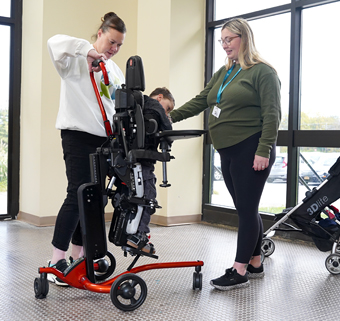 A boy stands in a Rifton Stander with the help of two therapists.