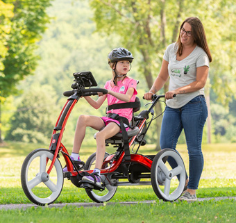 A girl rides on a Rifton Adaptive Tricycle while her caretaker steers from behind.