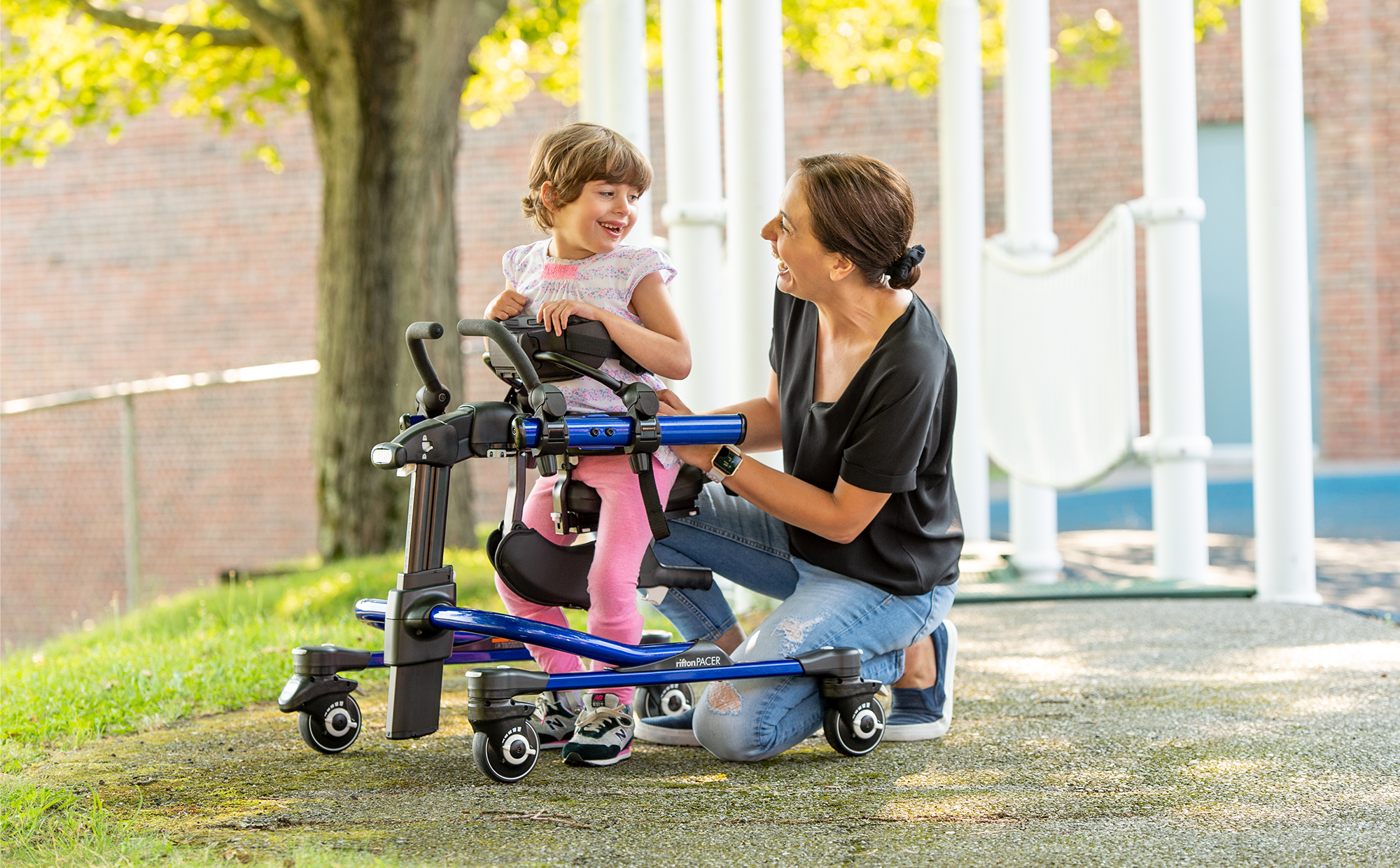 A little girl stands in a Rifton Stander and smiles at her caretaker.