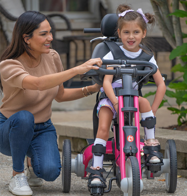 A girl riding on a Rifton Adaptive Tricycle with the help of her mother.