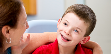 A boy sits on a Rifton HTS (Hygiene & Toileting System) while his caregiver smiles at him.