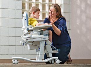 A boy sits on a Rifton HTS (Hygiene & Toileting System) while his caregiver smiles at him.