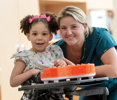 A girl standing in a Rifton Stander with therapist.