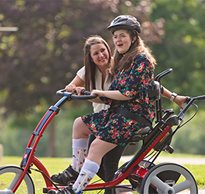A young lady rides on a Rifton Adaptive Tricycle, while her therapist walks beside her.