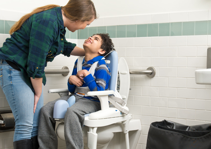 A boy sits on a Rifton HTS and smiles at his caretaker.