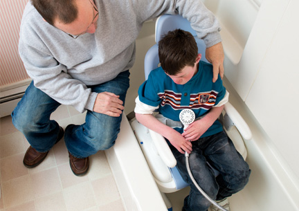 A boy sits in a Rifton HTS chair in a bathtub and talks with his caretaker.
